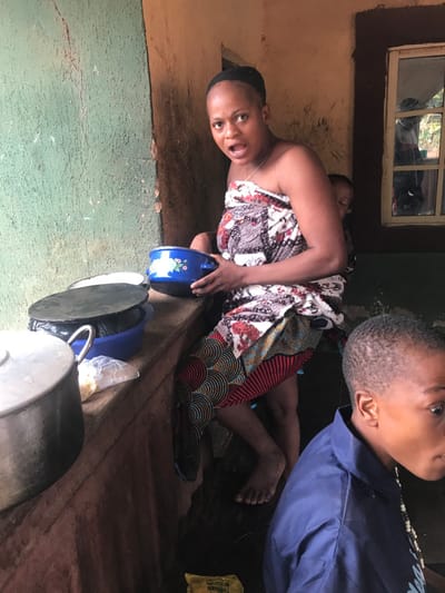 This image depicts a domestic scene in Ogodo, Nigeria. The primary subject is an adult female, likely a mother, positioned centrally. She has a medium complexion, dark hair pulled back, and is wearing a multi-patterned fabric wrapper that covers her torso and lower body. Her mouth is open as if speaking or reacting. She is holding a blue enamel bowl with a white floral design in both hands. A baby is secured to her back with a plain fabric wrap, partially visible over her right shoulder. The woman stands barefoot on a dark, possibly wet or muddy, floor.
To her right and in the foreground, a young child with dark hair and complexion is partially visible, wearing a blue collared shirt. The child is looking towards the right side of the frame.
To the left of the woman, on an elevated, rough-surfaced, earthy-toned counter or ledge, several cooking utensils are visible. These include a large silver pot, two dark-colored pots or lids, and a blue plastic bowl. A clear plastic bag containing white granular material is also present on the counter. The wall behind the counter is painted a faded teal-green with visible cracks and wear.
The wall to the right of the woman is an earthy brown color, appearing rough-textured. A rectangular window with multiple panes, framed in a darker color, is set into this wall, offering a blurred view of an outdoor environment. The overall lighting suggests an indoor or semi-enclosed space, illuminated by natural light. A piece of yellow packaging material lies on the dark floor in the lower-right quadrant.