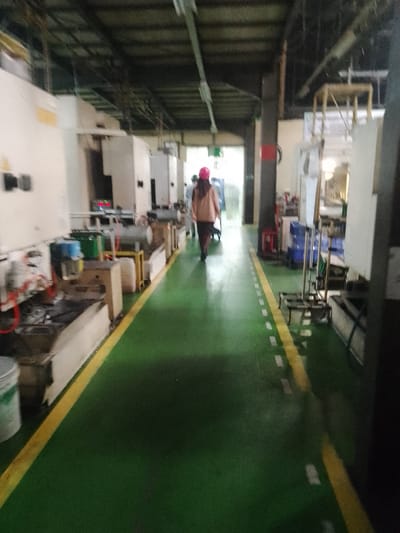 The image shows an interior view of a factory floor. A person wearing a pink hard hat and a light-colored jacket is walking away from the camera down a green floor with a yellow line marking the walkway. On either side of the walkway are various pieces of industrial machinery and equipment. The factory appears to be well-lit, with overhead lights and natural light coming from the end of the hall. The overall impression is one of a working industrial environment. This image was taken in Ciantra, Indonesia.