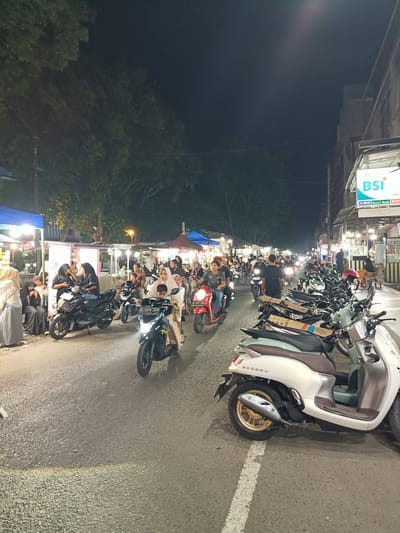 The image captures a lively nighttime scene in Bandarbireuen, Indonesia, likely during a street market or bustling evening event. A prominent element is the presence of numerous motorcycles and scooters navigating the road, indicating a busy atmosphere. Several individuals are visible, many of whom appear to be riding or walking around the various stalls.
The setting features a street lined with food and merchandise stalls, illuminated by bright lights, creating a vibrant and somewhat chaotic ambiance. Some stalls have blue and red awnings. People are dressed casually, and many women are wearing hijabs. The general mood seems to be one of activity and commerce.
Notable details include the time of day, which is clearly night, as evidenced by the artificial lighting and the dark sky. The road surface is asphalt, with a painted white line indicating a lane. The weather appears to be clear, allowing for the outdoor event to take place.
Visible text includes "BSI" in large, bright blue letters on a sign, likely for Bank Syariah Indonesia, positioned on a building facade along the street. There are also smaller text elements on some stalls and motorcycle license plates, but they are not clearly legible. The image conveys a sense of a typical Indonesian evening street scene, full of life and local activity.