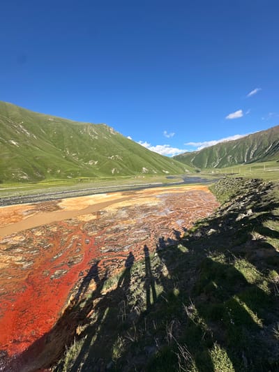 The image depicts a scenic view of a riverbed in Ketrisi, Georgia, characterized by its unique coloration. The river water exhibits a striking reddish-orange hue in some sections, contrasting with the surrounding earthy tones and the brown of the main water channel. These vibrant colors likely stem from mineral deposits or algae growth. Steep, lush green hills flank the river, their slopes covered in grass and showing signs of natural erosion with lighter patches of exposed earth. A clear blue sky with a few scattered white clouds dominates the upper portion of the image, suggesting fair weather. In the foreground, the shadows of several individuals are cast across the grassy bank and into the riverbed, indicating their presence and perspective. The overall environment suggests a natural, undeveloped landscape, typical of mountainous regions.