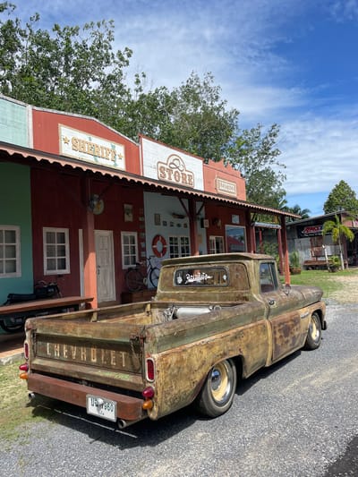 The scene is set outdoors in Ban Yid, Thailand, under a clear blue sky with scattered white clouds. A heavily rusted, lowered vintage Chevrolet pickup truck, displaying a green and brown patina, is parked on a light grey gravel surface, oriented with its rear facing the viewer. The truck features a custom wooden rear bumper, dual exhaust pipes, and a Thai license plate reading "บธ 1960 นราธิวาส." Its wheels have yellow/cream rims with chrome hubcaps and white sidewall tires. Cylindrical tanks are visible in the truck's open bed. A sticker on the rear window reads "จะอเมริกา."

In the background, a row of Western-themed buildings with weathered facades in red, light green, and light blue stand behind a covered porch area. Signs above the buildings read "Sheriff," "Store," and "Leather." Details on the buildings include white-framed windows, a white door, an exterior lamp, and a life preserver hanging on a blue wall. A motorcycle rests on a wooden bench to the left of the buildings, and a bicycle is parked against the building to the right. Further to the right and rear, a corrugated metal building featuring an "AMS The First in Speed" banner is visible, surrounded by palm trees and other green foliage. The scene is illuminated by bright daylight, casting distinct shadows. No individuals are present in the image.