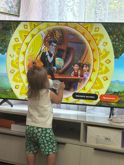 The image shows a young child, seen from behind, holding a bottle and watching a television screen. The television displays a colorful animated scene with characters and a decorative, circular pattern. Two buttons are visible on the screen, one labeled "Смотреть заставку" (Watch intro) and the other "Пропустить" (Skip). The child is wearing a white tank top with a floral pattern on the shorts. The television is placed on a white furniture unit. In the background, a window with sheer curtains is visible, and to the left, a framed picture hangs on the wall. The scene appears to be indoors.