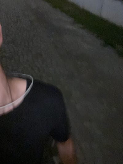 The image shows a person wearing a black t-shirt and a light-colored collar, possibly a necklace or part of the shirt. The person's neck and shoulder are visible, with skin tone appearing fair. The background is blurry and appears to be an outdoor environment with a dark, textured surface that could be asphalt or a similar ground covering. A thin strip of green vegetation is visible along the right side of the image, suggesting a roadside or park area. The lighting suggests it is either dusk or nighttime. No specific landmarks or identifying features of Gorna Malina, Bulgaria are present.