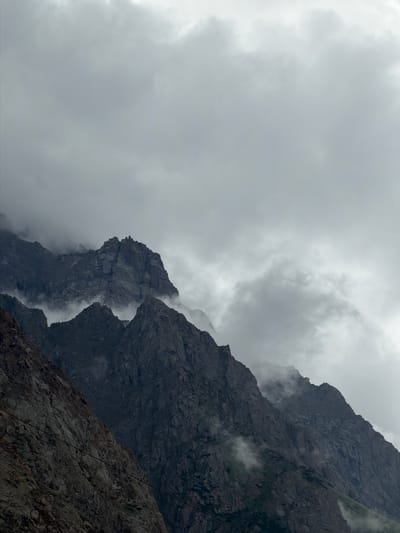 The image depicts a dramatic, overcast sky over a rugged mountain range. The sky is filled with thick, grey clouds, suggesting inclement weather or a perpetually cloudy atmosphere. Below the clouds, sharp, rocky peaks dominate the scene. These mountains are steep and jagged, with visible layers of rock and scree. Wisps of fog or mist cling to the slopes, partially obscuring some of the lower sections and adding to the atmospheric and somewhat mysterious mood. The overall color palette is muted, consisting primarily of grays, dark blues, and browns, characteristic of a high-altitude, cloudy environment. There are no people, animals, or man-made structures visible in the frame, making it a pure landscape shot emphasizing the raw, untamed nature of the mountains and the sky.