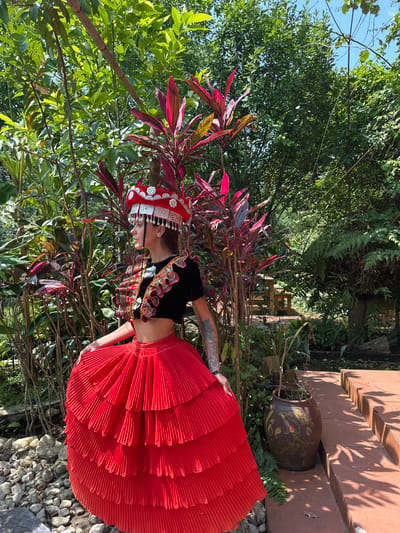 The image, taken in Thông Nguyên Commune, Vietnam, displays a person wearing elaborate traditional attire in a lush, sunlit outdoor setting. The individual, seen in profile, stands looking downwards and slightly to their left, appearing to pose for a photograph.

The attire consists of a black, short-sleeved top richly adorned with intricate, colorful embroidery and embellishments across the chest and sleeves. A vibrant, tiered, pleated, bright red skirt extends from the waist. A prominent, ornate silver-colored headpiece, intricately patterned and featuring dangling elements, adorns their head, with a single braid visible. Tattoos are visible on the person's left arm.

The scene is set in a verdant garden or natural area, characterized by dense green foliage, including trees and ferns, filling much of the background. Plants with striking red and burgundy leaves are prominently positioned around the subject. To the right of the person, a paved area with reddish-brown tiles is visible, featuring a large, ornate brown ceramic pot with gold designs. Clear shadows of leaves are cast upon this tiled surface, indicating bright daylight. The foreground features ground covered with rocks and pebbles. A clear blue sky is visible through gaps in the tree canopy. No text is visible in the image.