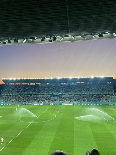 This image depicts a football (soccer) match or interval at Estadio Nuevo Arcángel in Córdoba, Spain, during dusk or sunset. The sky transitions from a deep purple overhead to orange at the horizon, with stadium floodlights fully illuminated, casting a bright glow over the scene. A faint airplane contrail is visible in the upper sky.

The perspective is from the stadium stands, with the tops of two heads visible in the extreme bottom foreground. Overhead, the dark, corrugated underside of the stadium roof is visible, with numerous bright floodlights affixed to its structure.

The stadium's multiple tiers of green seating are moderately populated with spectators. A large digital scoreboard in the distance displays "CÓRDOBA CF 1 45:00 0 CD MIRANDÉS," indicating a score of 1-0 with 45 minutes elapsed. Various sponsor advertisements line the perimeter of the pitch.

The well-maintained green football pitch with white line markings is being actively watered by four rotating sprinklers, which generate visible arcs and sprays of water. Several individuals are present on the pitch: one player in a white kit walks near the center circle, another in white is jogging in the lower-left foreground, and two players in darker kits (possibly red and black) are positioned near the far goal area. Distant hills are faintly visible beyond the stadium structure on the horizon.