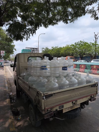 A flatbed truck is parked on the side of a street, laden with numerous large plastic water bottles, many of which appear to be full. The truck is tan and shows signs of wear. The water bottles are clear and stacked tightly in the truck bed, secured by a rope. The scene is set in Hyderabad, India, indicated by the license plate "TS09UD7396" on the rear of the truck, where "TS" is the state code for Telangana, the state in which Hyderabad is located. A sign with a speed limit symbol and "School Ahead" is visible on the left side of the road. The street is paved, with a sidewalk. Trees with dense foliage overhang the street and truck. In the background, construction is underway on a building, and there is traffic on the road, including cars and a motorcycle. The sky is cloudy.