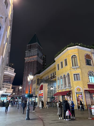 The image depicts a nighttime scene within a pedestrianized outdoor shopping and entertainment area in Esenler, Turkey, specifically characterized by architecture mimicking Venice, Italy.

Dominating the background is a tall, reddish-brown tower with a green pyramidal roof, resembling St. Mark's Campanile. This tower features tiered stone-colored sections near its base and top, with multiple arched openings on the upper levels. To the right of the tower stands a multi-story building with a yellow stucco facade, adorned with numerous arched and rectangular windows, some featuring decorative brickwork. The roofline of this yellow building is topped with green foliage. Below, red awnings cover storefronts, and a glass-enclosed structure provides an entrance or walkway between sections of the building. Visible shop signs include "CARS PARK" and "FLO." On the far left, a lighter-colored building with classical architectural elements and balustraded balconies is partially visible.

The ground is paved with dark gray interlocking stones, forming a wide pathway. Several ornate, multi-lamp streetlights illuminate the area, positioned centrally within the pedestrian zone. Numerous individuals, estimated between 20 and 30, populate the scene. They are seen walking, standing in small groups, and congregating. In the lower right foreground, a group of four individuals is visible, one holding a clear umbrella, and another carrying shopping bags. People are dressed in casual evening attire, including jackets and coats. The sky above is dark, indicating night, with artificial light sources providing illumination throughout the environment, highlighting the buildings and the pathway.