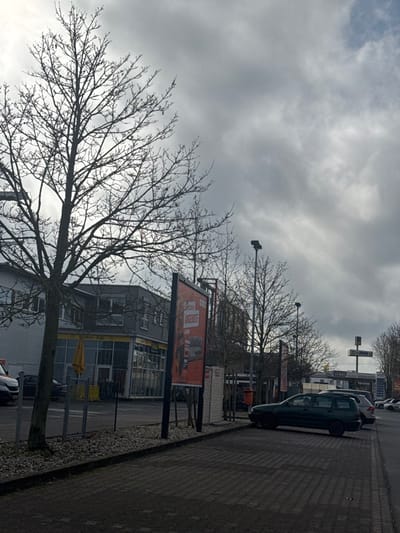This image captures an outdoor commercial environment under an overcast sky in Gießen, Germany, specifically at Pistorstraße 15, 35394 Gießen. The scene is characterized by bare deciduous trees, suggesting late winter or early spring.
In the foreground, a paved parking area, composed of dark interlocking pavers, extends from the right. A curb and a gravel strip, populated by multiple bare trees and several tall streetlights, separate the parking area from the buildings. A dark green station wagon is parked diagonally in the foreground, facing towards the left. Behind it, a silver station wagon is also parked. A dark red cylindrical bin is partially visible behind one of the billboards.
To the left, a two-story building features "Liebmann Cafe" on its ground floor, with large windows, a yellow outdoor umbrella, and yellow safety barriers. Adjacent to the cafe and within the gravel strip, a large vertical billboard displays red text "ANGEBOTE" (offers) and images of furniture, with the "OBI" logo visible at the top.
Further into the midground, additional commercial structures are visible, including a prominent sign indicating "TOTAL" and "HÖSEL," which appears to be a gas station or service area. Multiple other vehicles are parked further down the street. No individuals are discernible within the frame.