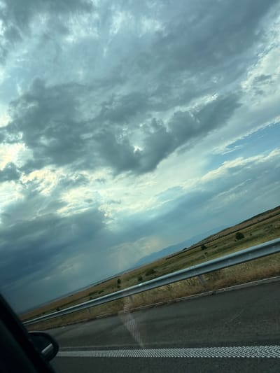 This is a low-angle, rotated, full shot of a highway in Zheleznik, Bulgaria. The perspective is from inside a vehicle, looking out. The scene is dominated by a dramatic sky filled with varied clouds. The clouds range from dark, ominous grey masses to lighter, wispy formations, with patches of bright sunlight breaking through in places, suggesting a dynamic and possibly changeable weather pattern. The sky occupies the majority of the frame, creating a sense of expanse and depth.

To the right of the sky is the highway itself. The asphalt road is visible, with a clear white dashed line marking the lane. The shoulder of the road is visible, bordered by a metal guardrail. Beyond the guardrail is a strip of dry, yellowish-green vegetation, indicating a dry or arid environment, with a few sparse trees dotting the landscape. The ground appears flat and stretches into the distance.

The time of day is suggested by the lighting of the clouds and the overall brightness of the scene, likely late afternoon or early evening, given the dramatic interplay of light and shadow in the sky. The overall mood is somewhat contemplative or perhaps a little unsettled due to the dramatic sky. There is no visible text in the image.
