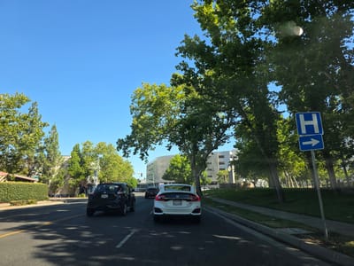 The image depicts a scene from a vehicle on a sunny day, likely in Sacramento, California. The sky is a clear, bright blue, suggesting good weather. The setting is a suburban or commercial area with mature trees lining the street.
In the foreground, a white Honda Civic is driving in the right lane, with its rear visible. Ahead of it, a black Nissan Juke is also driving in the same lane. To the left, another vehicle, partially obscured, is visible in the left lane. The road is paved with asphalt and has clear lane markings, including yellow lines separating opposing traffic and white lines marking the lanes.
On the right side of the road, a sidewalk runs alongside a grassy area with several large trees. A blue sign with a white "H" symbol and a white arrow pointing to the right is mounted on a pole. Further down the street, a modern building with a curved, white facade is visible, appearing to be a commercial or institutional structure. A sign at the base of a monument reads "Sutter's Gardens."
The time of day appears to be late morning or early afternoon, given the bright sunlight and the shadows cast by the trees. The overall mood is peaceful and ordinary, depicting a typical day of driving in a well-maintained urban environment. No people are clearly discernible in the image, as the focus is on the road and surrounding environment.