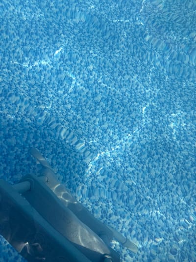 The image shows the patterned floor of a swimming pool, with sunlight refracting through the water, creating rippling light effects. A portion of a pool ladder is visible in the lower left corner. The scene appears to be indoors or shaded, as indicated by the controlled lighting and the lack of direct sunlight or shadows on the pool deck. There is no indication of people or any other activity. The location in the city of Dzerzhinsk, Russia, is not discernible from the visual information provided in the image.
