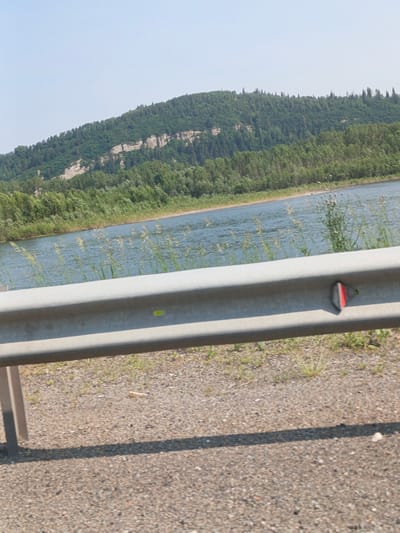The image captures a scenic view of a wide, calm river flowing alongside a tree-covered hillside. In the foreground, a metal guardrail runs horizontally across the frame, with a red and silver reflector attached to its right side. The ground in front of the guardrail is covered in gravel and sparse green vegetation. The river reflects the clear, light blue sky, and the opposite bank is densely populated with various types of trees. The hillside rises steeply from the river, adorned with a mix of deciduous and coniferous trees, and a distinct rocky ledge is visible partway up the slope. The overall impression is one of a tranquil natural landscape, likely located outside of an urban area. Given the reference to Bezrukovo, Russia, this scene is likely situated within the natural surroundings of that region.