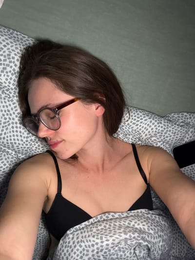 A woman with glasses lies on a bed. She is wearing a black bra. A smartphone is visible above her head. The bedding is patterned in gray and white. The background includes a solid light green surface. No information is available about the city of Kranj, Slovenia.