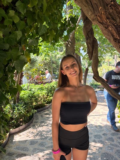 A photograph depicts a young woman in a black tube top and black shorts standing outdoors. She is smiling and looking directly at the camera. A tree branch is positioned above her head. In the background, another person wearing a grey shirt and jeans is standing on a stone path. Further back, trees and vegetation are visible, and an older person is seated on a bench. The photo appears to be taken in a natural setting within Bunyola, Spain, based on the scenery and context.