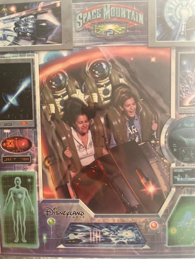 The image is a photograph of a souvenir from Disneyland Paris, featuring a ride called Space Mountain: Mission 2. The central part of the image shows two women on the ride, wearing spacesuits and helmets, with their mouths open in excitement. The background depicts a fiery, space-themed landscape with asteroids and a bright star. Surrounding this central image are various graphic elements, including a spaceship, a diagram of a human body, a radar screen, and the Disneyland Paris logo. There is no indication of the city of Badalona, Spain, within the image or its context.