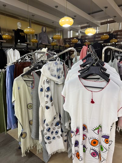 The image shows a clothing store in Acıpayam, Turkey, with racks of clothes displayed. Several mannequins are visible in the background, some wearing hoodies and t-shirts. Hanging light fixtures with yellow shades illuminate the store. The foreground features a variety of garments, including dresses and tops, on hangers. Some of the clothing has colorful patterns, such as eyes and floral designs. The store appears to be well-stocked and organized.