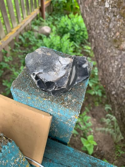 A dark, angular rock with lighter grey inclusions sits on a weathered, turquoise-painted wooden surface, possibly part of a bench or railing, in an outdoor setting. The rock appears to be a piece of flint or obsidian, with sharp edges and a partially polished surface. The wooden surface shows signs of age and wear, with chipping paint and some yellow and white speckled discoloration. In the background, a weathered wooden fence with a horizontal yellow beam is visible, along with lush green vegetation and a tree trunk. The ground is covered with soil and more greenery. The overall impression is of a natural scene with a man-made element, likely in a rural or garden environment within the Орлеўскі сельскі Савет, Belarus.