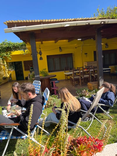 Outdoors in Cordoba, Argentina, four people sit in folding chairs on a grassy lawn near a yellow building. One person in a blue jacket sits upright. A person with long blonde hair is partially obscured. A person wearing black mixes food in a bowl held by another person. The building has a brown roof and a covered porch with visible tables and chairs. Decorative elements hang on the wall. Sunlight illuminates the scene from the left, casting shadows. In the foreground, foliage with red and white blossoms frames the scene.