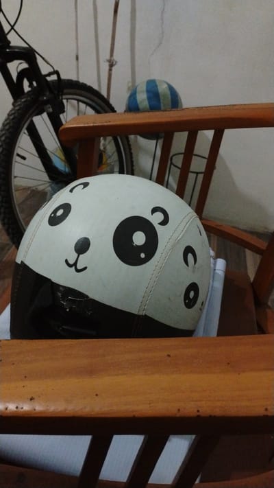 In Banjarsari, Indonesia, a white helmet with a cartoon panda face design sits on a wooden chair. The helmet is positioned towards the left side of the frame, with its black lower section partially obscured. To the left of the helmet, a portion of a bicycle is visible, including its front wheel and black frame. In the background, a blue and green striped ball rests behind the chair, and a white wall with some vertical wooden elements is present. The lighting suggests an indoor setting.