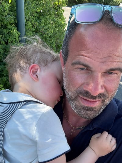 A medium-shot, candid photograph depicts a Caucasian man embracing a young child against a background of lush greenery, possibly a garden or park setting. The man, with fair skin, short dark hair, and a salt-and-pepper beard, is wearing a dark blue top and has sunglasses perched on his head. He is looking down at the child with a tender expression. A gold cross necklace is visible around his neck. The child, fair-skinned with light blonde hair, is lying against the man's chest, seemingly asleep. The child is wearing a light-colored polo shirt with navy blue striped suspenders. The setting includes green foliage, suggesting an outdoor environment, possibly a park or garden. Considering the location is Bredene, Belgium, the setting could be a local park or residential garden within the city. There is a green metal fence or railing present on the left side of the photo.