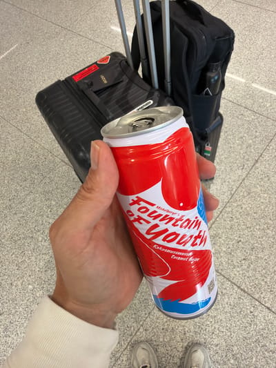 A person is holding an open can of "Fountain of Youth Coconut Water" while standing in an airport terminal. The can has a red and white design with blue accents at the bottom. In the background, a black rolling suitcase and a black backpack are visible, along with part of a suitcase handle. The floor is tiled with a speckled grey pattern. The image is taken from a first-person perspective.