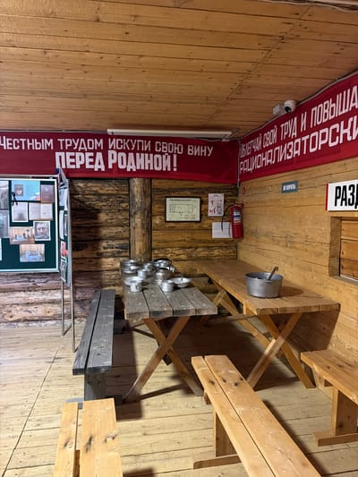 The image depicts an indoor scene, likely a museum or historical exhibit, with rustic wooden walls and ceiling. A prominent red banner stretches across the top, bearing white text in Russian. To the left, a display board features photographs and documents. In the center, wooden tables and benches are set up, with several metal pots and utensils on the tables. A fire extinguisher is mounted on the wall to the right, near a sign with Russian text. The overall atmosphere suggests a historical setting related to work or military life.
The location is specified as Ust'-Pyzas, Russia. This detail is crucial for contextualizing the historical significance of the exhibit. Given the imagery and the text on the banner, the exhibit likely pertains to Soviet-era history, possibly related to labor camps, industrialization, or wartime experiences. The rustic wooden construction and the utilitarian furnishings are consistent with representations of such historical periods in rural or remote regions of Russia. The presence of what appears to be personal photographs and documents on the display board suggests an attempt to humanize the historical narrative. The metalware on the tables could represent communal dining or basic provisions typical of the era being depicted. The fire extinguisher and the "Do Not Smoke" sign are standard safety features in public spaces, but their placement within this historical exhibit adds to the sense of realism.