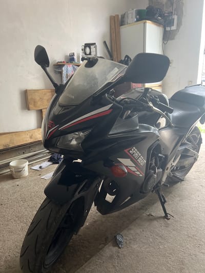 The image shows a black Honda motorcycle parked indoors, likely in a garage or storage area. The motorcycle has a "Spirit Beast" sticker on its side. A GoPro camera is attached to the front fairing. Various objects are scattered around, including wooden planks, a white cup, and miscellaneous items on a shelf. The environment appears cluttered and utilitarian. While the image was taken in Cherliany, Ukraine, there is no specific indication of the location in the city.