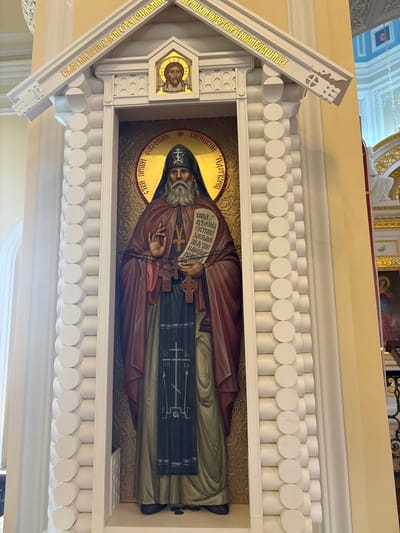 A religious icon featuring a full-length depiction of a bearded elder monk is displayed within a white, log-cabin-style shrine. The monk, dressed in red and black robes with a dark hood, holds a scroll in his left hand and blesses with his right. Above him, in a golden halo, is an image of Jesus Christ. The shrine is adorned with gold Cyrillic lettering on its roof, which is partially visible. The background is a pale yellow wall. The icon is located within a church, as suggested by the architectural elements and other religious iconography visible in the background, including a depiction of Mary and Jesus and ornate gold details. Based on the context of the lettering and the architectural style, this icon is likely located in the Church of the Holy Trinity in Gatchina, Russia.