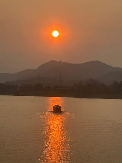 The scene depicts a sunset or sunrise over the Perfume River in Kim Long Ward, Vietnam. A bright, spherical orange sun is centrally positioned in the upper portion of a hazy, orange-toned sky. Below the horizon, a series of dark, undulating mountains are silhouetted against the sky. On the far bank of the river, between the mountains and the water, a line of silhouetted trees and indistinct man-made structures, including what appear to be two transmission towers, are visible. The wide river occupies the lower two-thirds of the image, reflecting the sun's intense orange light as a prominent, shimmering vertical streak. A small, dark-colored, enclosed boat with a cabin is positioned centrally within this sun reflection on the water, moving away from the viewer and leaving a subtle V-shaped wake. No individuals are discernible on the boat or within the wider environment.
