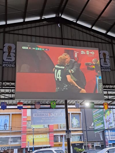 The image shows a large outdoor screen displaying a soccer match at the FIFA World Cup 2026. The screen shows a score of Canada 4, Qatar 0 at 71:35 minutes. Two men are visible on the screen; one is wearing a black jersey with the number 14 and the other is in a suit. Below the screen, several national flags are hanging, including those of Australia, Paraguay, the USA, and Brazil. In the background, there are buildings with various signs, including "guardian" and "Qyas," indicating a commercial area. The overall scene suggests a public viewing of the soccer match in a city environment, likely Bireuen, Indonesia, given the presence of Indonesian script on some of the banners and the style of the buildings.