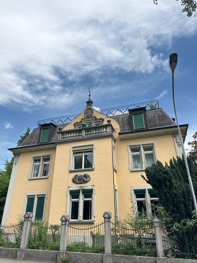 This media file displays a close-up, rotated view of a yellow building in Zurich, Switzerland. The building appears to be an older architectural style, featuring decorative elements above the windows and along the roofline. There are several windows visible, some with green shutters. A wrought-iron fence and stone pillars are in the foreground, with lush green foliage partially obscuring them. A large evergreen tree is on the right side of the building. The sky is a bright blue with scattered white clouds, suggesting it is daytime with fair weather. A lamppost is visible in the upper left quadrant of the image, extending towards the building. The image is oriented vertically, with the top of the building appearing on the left and the sky on the right. No people or specific activities are depicted, but the scene conveys a sense of quiet residential or commercial property. No text is visible in the image.