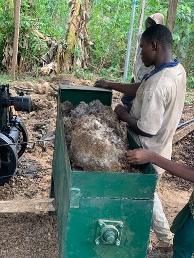 The image shows two men in Egbeda, Nigeria, at the coordinates 7.12 latitude, 5.362 longitude, loading a large, green, metal hopper with a mass of brown fibrous material. The man closest to the camera is leaning into the hopper with his left hand holding a piece of the material while his right hand is resting on the top of the hopper. The other man, partially obscured, is standing behind him, looking at the material in the hopper. The hopper is attached to a dark-colored, industrial machine that is visible in the lower portion of the image. The ground surrounding the machine and the hopper is covered in a layer of dry, brown, fibrous material. Thick vegetation fills the background. The image appears to show a process of agricultural production, with the fibrous material likely being a type of crop residue or fiber that is being processed by the industrial machine.