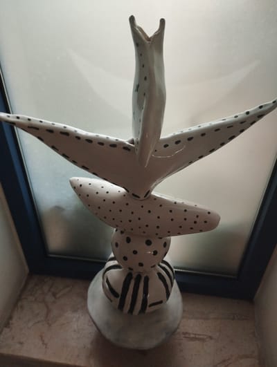 A tall, abstract ceramic sculpture, primarily white with black polka dots and stripes, stands on a marble-like surface against a blurry window. The sculpture has a layered design, featuring spread wings, a central bulbous section, and a striped base. The top of the sculpture is open, resembling a beak or mouth. The environment appears to be indoors, near a window with diffused light. The location is not specified in the image itself.