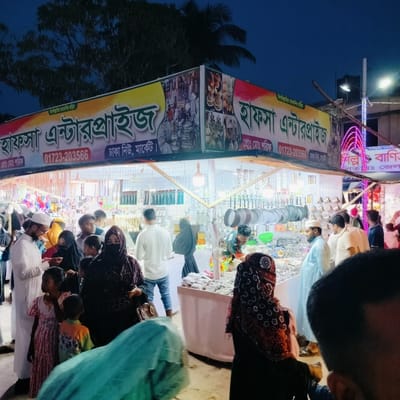 This nighttime image captures a bustling market stall, likely in Charfasson, Bangladesh, given the context. The scene is illuminated by artificial lights, indicating evening or night hours, with a dark blue sky and silhouetted trees in the background.

The main subject is a large, brightly lit market stall named "Hafsa Enterprise" (হাফসা এন্টারপ্রাইজ). The stall features various household items, kitchenware like pots and pans, and possibly jewelry or decorative pieces, displayed on tables and hanging from racks. Numerous people, including men, women, and children, are seen browsing and interacting within and around the stall. Many of the women wear hijabs or head coverings, and some men wear prayer caps (taqiyahs), reflecting local cultural attire. The atmosphere appears lively and crowded.

Visible text on the main banner prominently displays "বিসমিল্লাহির রাহমানির রাহিম" (In the name of Allah, the Most Gracious, the Most Merciful) at the top. The enterprise name "হাফসা এন্টারপ্রাইজ" (Hafsa Enterprise) is repeated. Below it, the contact number "01723-203566" and location "ঢাকা নিউ, মার্কেট" (Dhaka New Market) are also visible. Additionally, "প্রোঃ মাঃ শরিক" (Prop: Mr. Sharif) is listed as the proprietor. Another sign on the far right, partially obscured and decorated with neon pink lights, contains text like "শল্প বাণিত" (Industry/Crafts and Trade) and "গিতায়ঃ জেল" (possibly a district or area name). The people appear engaged in shopping, some examining goods closely, others simply moving through the crowd.