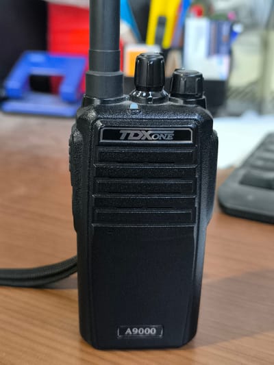 A black handheld radio, identified by the logo "TDXONE" and model number "A9000", is positioned on a wooden surface, possibly a desk. The radio has two knobs on top, with markings "3", "1", and "2" visible near one of the knobs. A black cord is attached to the side of the radio. In the blurred background, office supplies such as colored folders and what appears to be a keyboard can be observed. The scene is indoors.