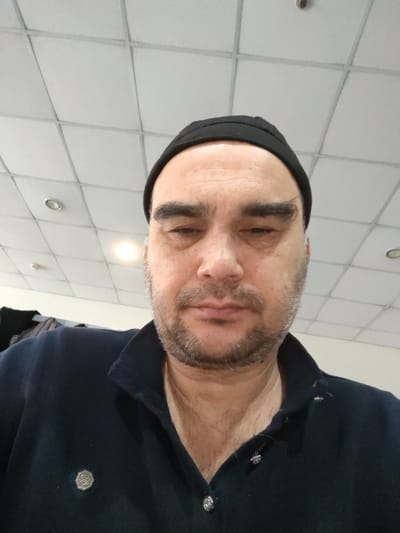 The image is a medium close-up selfie of a man in his 40s, with fair skin, dark hair, and a beard. He is wearing a black beanie and a dark blue collared shirt with silver buttons. The background appears to be an indoor setting with a tiled ceiling and lighting fixtures. There is no indication of the man's location within the city of Shchyolkovo, Russia. The man's expression is neutral, and he is looking directly at the camera. His facial features include dark, thick eyebrows, a prominent nose, and thin lips. His beard is short and appears to be lightly trimmed. The lighting in the image is bright, casting some shadows on his face.