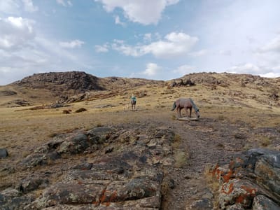 The image depicts a rocky, arid landscape under a sky with scattered clouds. In the foreground, large, weathered rocks with patches of orange and green lichen are visible. A dirt path, scattered with gravel, leads into the middle ground. On the right side of the path stands a life-sized statue of a horse, its head lowered as if grazing. Further into the middle ground, a person in a light blue top and tan pants is standing on the path, accompanied by a small, light-colored dog. The person appears to be looking towards the left. The background features rolling hills covered in dry grass and scattered rocks, with more rugged, rocky formations in the distance. The overall impression is of a remote, natural environment, possibly a hiking trail or park area. This scene is located in the vicinity of Мақаншы, Kazakhstan, an area known for its steppe and mountainous terrain.
