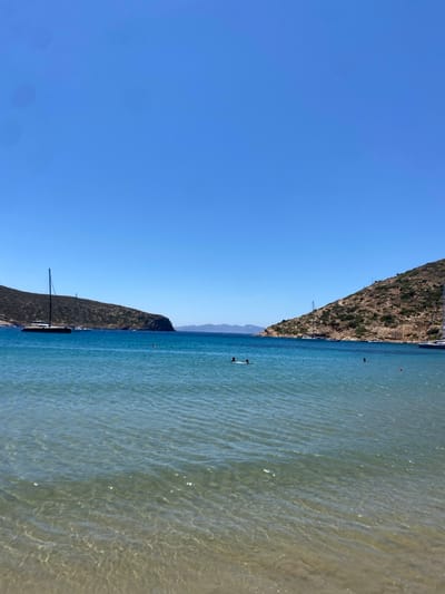 The image depicts a sunlit coastal bay identified as being in the vicinity of the city of Απολλωνία, Greece. The foreground features shallow, transparent turquoise water, revealing the sandy seabed with light reflections across its surface. In the mid-ground, the water transitions to a deeper blue. Two individuals are visible swimming, their heads just above the water line. To the left, a large, dark-hulled sailboat with a tall mast is anchored near a rocky, arid hill covered in sparse, scrubby vegetation. A similar rocky hill rises on the right side, partially obscuring another white sailboat moored near its base. These two landmasses frame a narrow strait that opens to the wider sea, where distant, hazy blue mountains are faintly visible on the far horizon. The sky overhead is uniformly clear and bright blue, devoid of clouds.