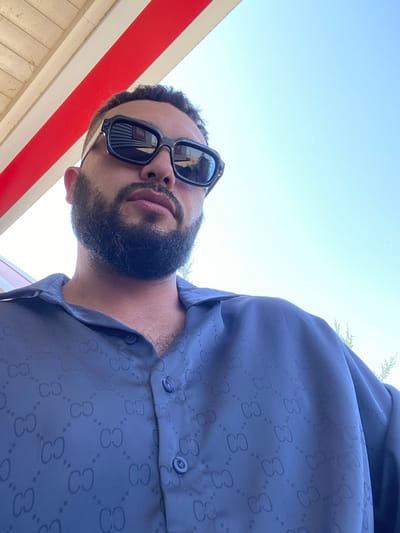 A man wearing sunglasses and a blue shirt with a repeating pattern is pictured. He has a beard and is looking away from the camera. The background consists of a blue sky and a red and white striped structure. The location is not identifiable within the city of Korkmaskala, Russia.