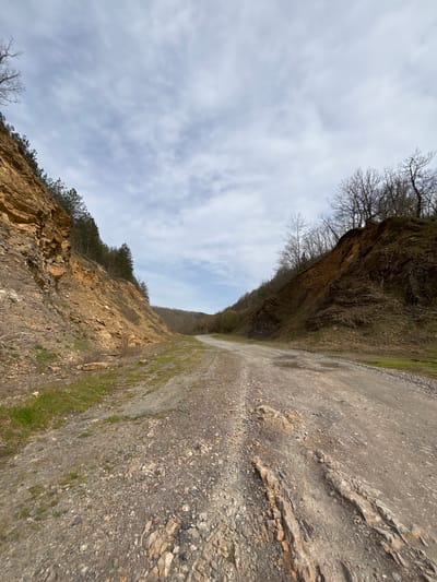 The image shows a gravel road or path flanked by rock cliffs or embankments on either side. The left embankment is darker, possibly damp or mossy, with bare trees near the top. The right embankment is lighter in color with some vegetation and small trees. Above the embankments, a cloudy sky is visible. There are no people or other obvious signs of civilization present. The image does not appear to be in Kosti, Bulgaria. The terrain looks more like a quarry or hillside path. There is nothing definitive to say that the picture is from a city.