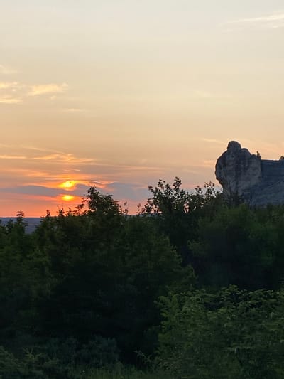 This is a vertical, medium shot photograph of a sunset over a rocky outcrop and a dense forest in Krasnomakskoye, Ukraine. The sky is a gradient of pale yellow at the top, transitioning to orange and pink hues near the horizon. A bright orange sun is visible, partially obscured by thin, wispy clouds. In the middle ground, a prominent, weathered rock formation with a distinctive, human-like silhouette stands on the right side of the frame. In front of the rock formation and stretching across the lower half of the image is a thick canopy of dark green trees and foliage, with some lighter green bushes in the foreground. The overall scene captures a serene and natural landscape at dusk.