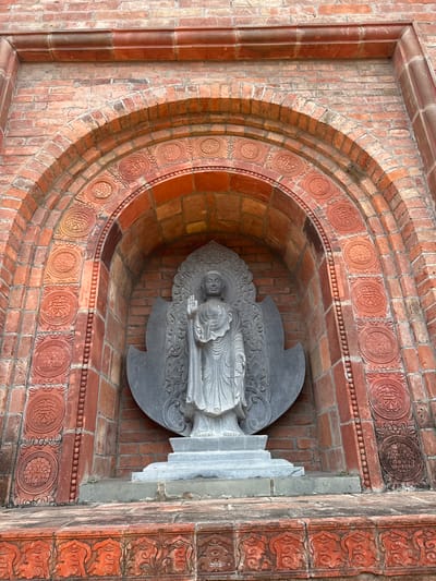 The image shows a Buddha statue standing in an alcove within a brick structure. The alcove is arched and decorated with circular motifs carved into the bricks. The Buddha statue is made of grey stone and is depicted standing with its right hand raised. The base of the statue is a tiered platform. The surrounding brickwork is aged and shows variations in color. The scene is outdoors, and the lighting suggests daytime. The location is identified as Tây Hoa Lư Ward, Vietnam.