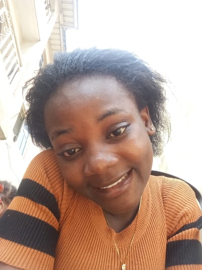 A female individual, appearing to be an adult of African descent, is depicted in a close-up, slightly low-angle shot, presumably taken in Agburu Uke, Nigeria. The subject possesses dark skin, dark, curly hair, and dark eyes, and is smiling, revealing her upper teeth. She is wearing an orange-brown ribbed long-sleeved top with two black horizontal stripes on the visible left arm. A thin gold-colored chain necklace with a small, light-colored pendant is visible around her neck, and a small, light-colored stud earring is present in her right earlobe. In the lower left background, a blurred portion of a second individual, consistent with a child exhibiting dark hair and a red accessory, is partially visible. The environment appears to be an outdoor or brightly lit area, with a light-colored building structure featuring windows and a white railing or grille visible in the upper left background. The upper right background is overexposed white, indicative of a bright sky or wall.