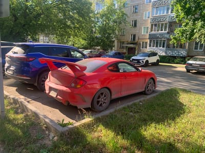 This image captures a sunny daytime scene on a city street in Ivanovo, Russia, likely in a residential area. In the foreground, a bright red sports car with a prominent rear spoiler is parked at an angle. The car appears somewhat dusty. Beside it, a blue SUV is also parked. Further down the street, a white SUV and a light-colored sedan are visible, along with other vehicles partially obscured. The backdrop features a multi-story apartment building with balconies and trees lining the street, suggesting a typical urban environment. The ground is paved with asphalt and there is a strip of grass and a concrete curb separating the parking area from the sidewalk. The overall atmosphere is calm and static, with no people or discernible activity.