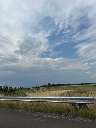 The image depicts a roadside scene in Malo Konare, Bulgaria, under a partly cloudy sky. The foreground is dominated by a silver metal guardrail running along the edge of an asphalt road. To the left of the guardrail is a grassy embankment with dry, tall grass and some scattered green foliage, suggesting a rural or natural setting. Beyond this, a vast field stretches into the distance, appearing to be agricultural land with some areas of green crops and others of dry, harvested stalks. A line of trees marks the far edge of the field, and further still, hazy hills are visible on the horizon. The sky above is a mix of bright blue patches and soft, wispy white clouds, indicating daytime, likely mid-morning or afternoon, with fair weather. The overall impression is one of a serene, open landscape. No people or text are visible in the image.