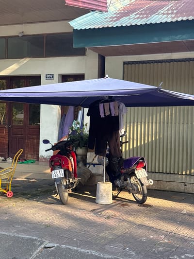 An outdoor daytime scene in Lao Cai Ward, Vietnam, depicts two motorbikes parked on a tiled sidewalk adjacent to a building. A large blue awning extends over the motorbikes and onto the sidewalk. Clothing is hanging on a rack near the motorbikes. A yellow shopping cart is also visible on the sidewalk. The building has a combination of corrugated metal and painted walls, with a door and windows visible.
