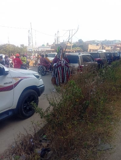 This image depicts a bustling street scene at Itakpe Junction, Nigeria, under bright daylight conditions. In the midground, a central figure is prominently featured, clad in an elaborate, full-body traditional costume characterized by vertical black, white, and red stripes on the lower half, and a complex, multi-layered upper section including a headpiece with green elements. This individual holds a thin, multi-branched stick or whip in their right hand.

To the immediate left in the foreground, the front quarter of a white Toyota Hilux pickup truck is visible, featuring black rims and a silver bumper. The paved road beyond the Hilux is occupied by numerous vehicles and people. Several motorcycles ("okadas") carrying riders and passengers are present in the midground, with one rider identifiable by a red shirt and orange helmet. A line of cars and SUVs extends into the background on the right, suggesting traffic or parked vehicles.

In the distant background, a prominent mosque with two tall, white minarets and a green dome serves as a distinct landmark. Other urban structures are visible behind the vehicles and the mosque. The immediate foreground on the right consists of a strip of roadside vegetation, appearing somewhat dry and sparse, with visible litter and debris mixed within. The sky is bright and largely overexposed.