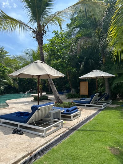 The image shows a poolside lounge area at a resort in Santa Teresa, Costa Rica. Two white umbrellas with white frames are positioned above two lounge chairs. The chairs are made of white metal frames and are covered in blue cushions. The chairs are situated on a paved patio, which is surrounded by lush green grass and tropical plants. There are multiple trees in the area, including a tall palm tree in the foreground. The ground is made of white concrete tiles with a drainage system in the background. The chairs are empty and facing the pool, which is visible in the background. The pool is surrounded by trees and lush foliage. The scene is serene and inviting, with the lush greenery and the tranquil ambiance suggesting a relaxing environment.