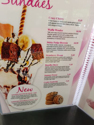 The image displays a menu featuring various ice cream sundaes, with the text written in English. The menu is likely found in a dessert shop or restaurant. The sundaes are described with ingredients such as gelato, whipped cream, sauces, fruits, and wafers, with prices ranging from 8.49 to 10.99. There are also images of sundaes and some of the ingredients like cookies. The overall presentation is bright and appealing, designed to entice customers. There is no information within the image to indicate its location in Bedford, United Kingdom, or any specific details about people or actions.